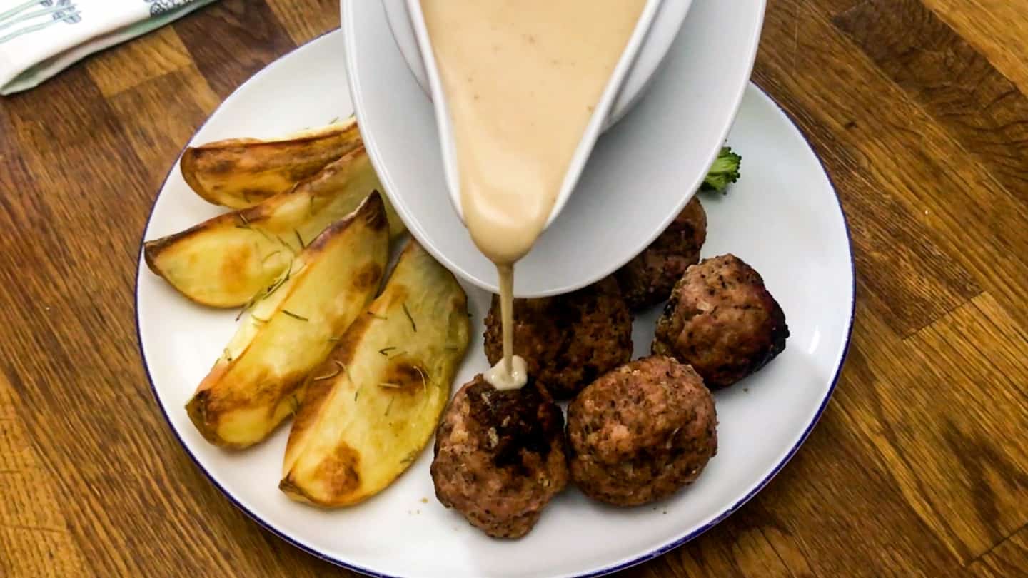 Gravy being poured from gravy boat on top of 5 perfectly browned meatballs. Served with a side of crispy potato wedges.
