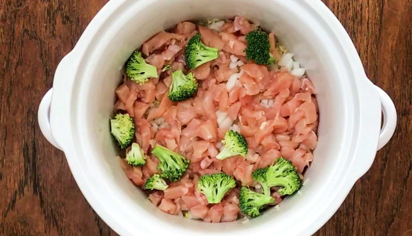 slow cooker pot with rice, onions, and garlic, chicken breasts and broccoli on top